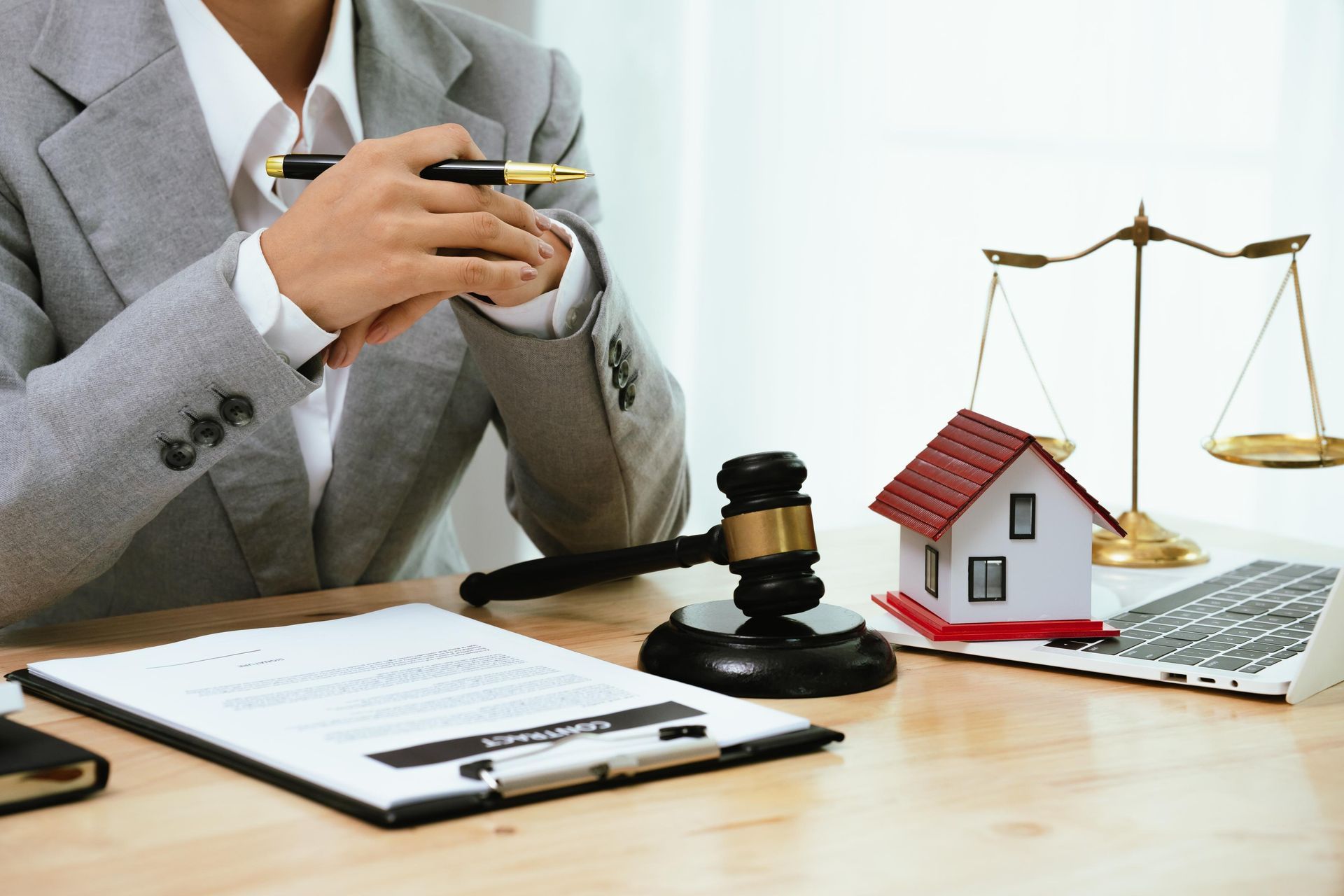 Lawyer's desk: person in a blazer with a pen, gavel, house model, and scales of justice.