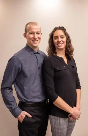 Two smiling coworkers standing indoors, wearing business attire against a plain wall.