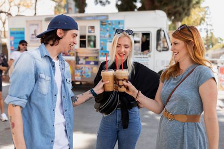Three friends clink iced coffee cups beside a white food truck outdoors.