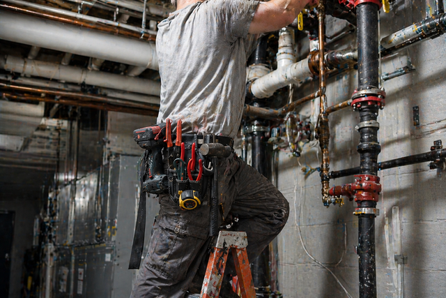 Technician on a ladder repairing exposed pipes in an industrial basement.