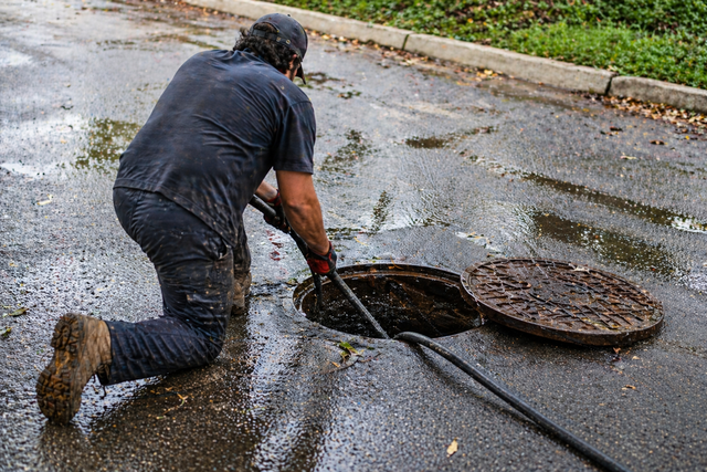 Worker kneeling on wet pavement unclogs a manhole with a long tool, water around the opening.