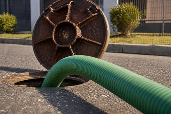 Green hose inserted into a manhole on a street, with the cover leaning against the curb.