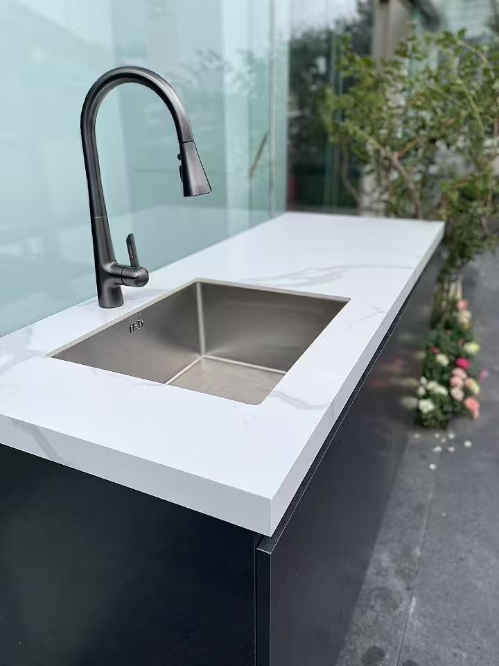 Modern outdoor kitchen sink with black faucet and white countertop near a glass wall