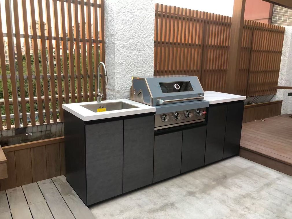 Outdoor kitchen with sink and stainless steel grill on a wooden deck, beside a slatted fence