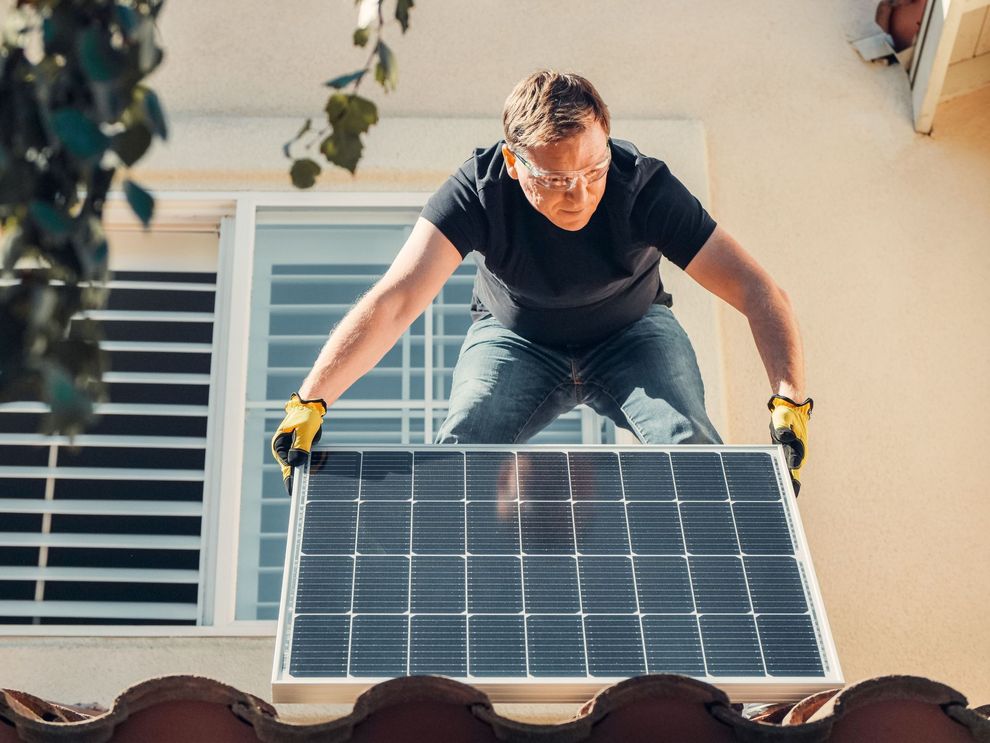 Man in gloves lifting a solar panel onto a rooftop beside house windows