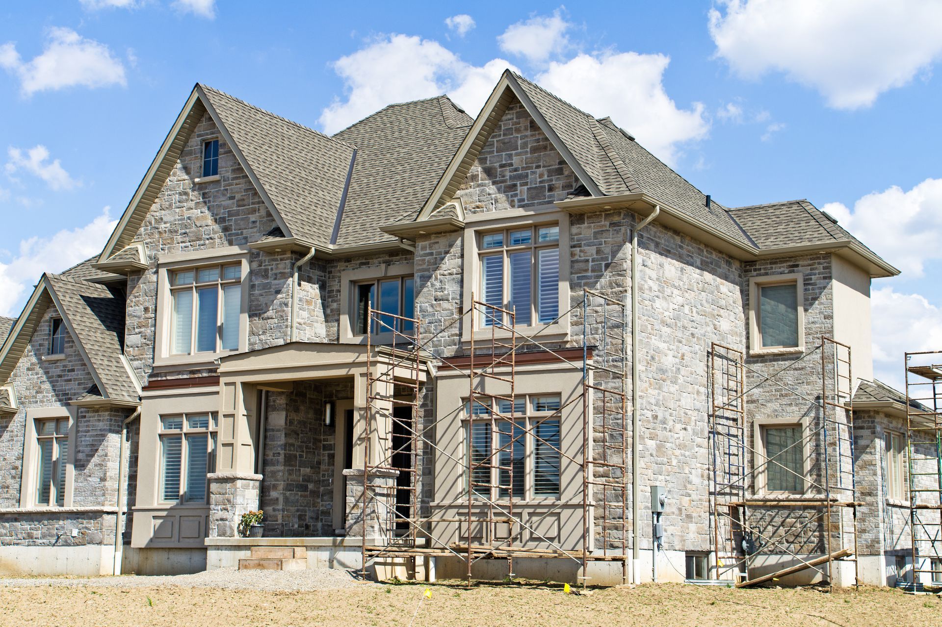 Two-story house with stone facade, under construction, against a blue sky with clouds.