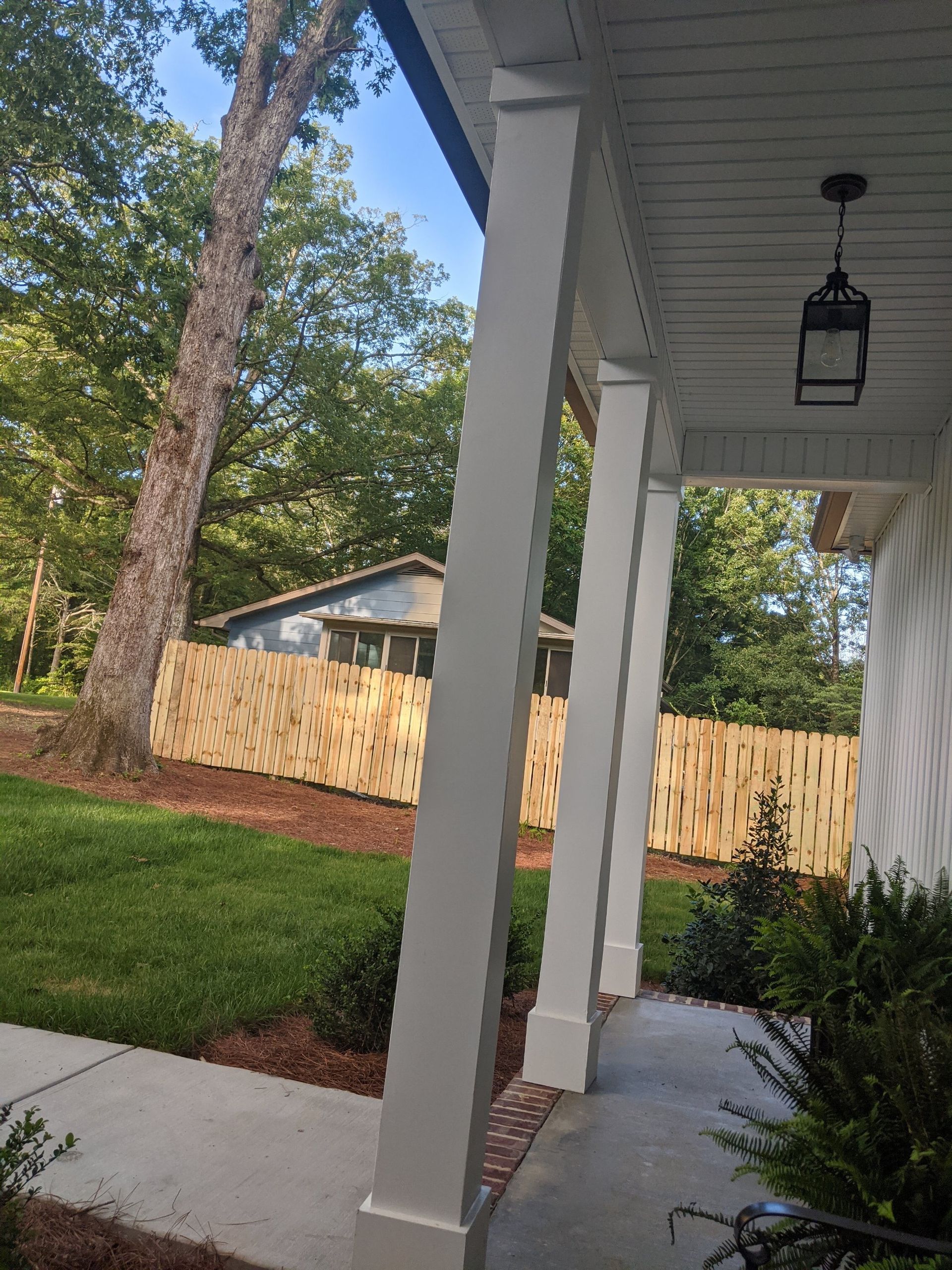 White porch columns with a hanging lantern, green grass, and a wooden fence in the background.
