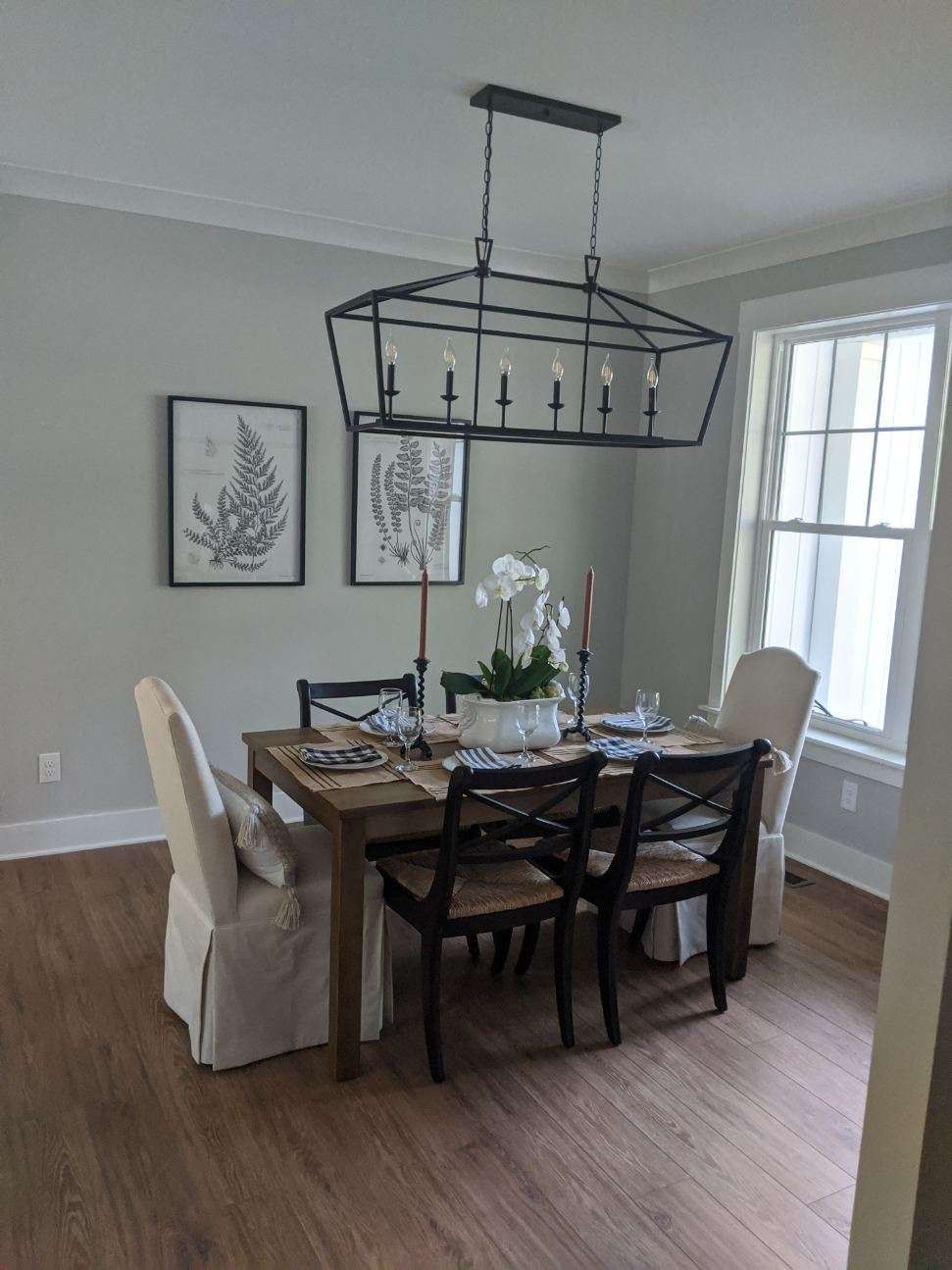 Dining room with table set for meal; dark chandelier above; two framed botanical prints on wall.
