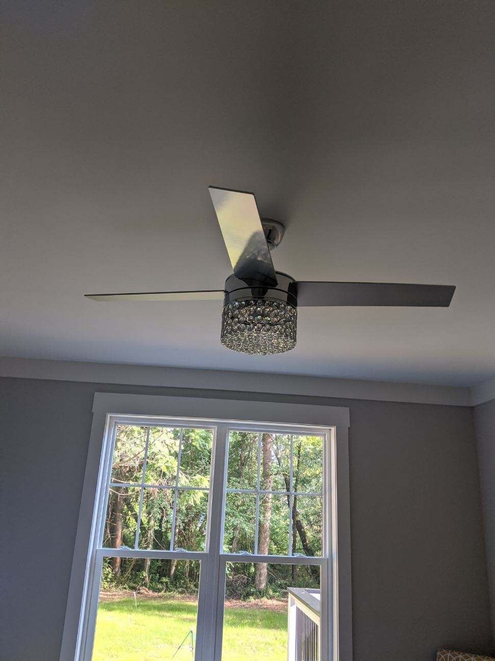 Ceiling fan with crystal light fixture, gray blades, in room with window overlooking trees.