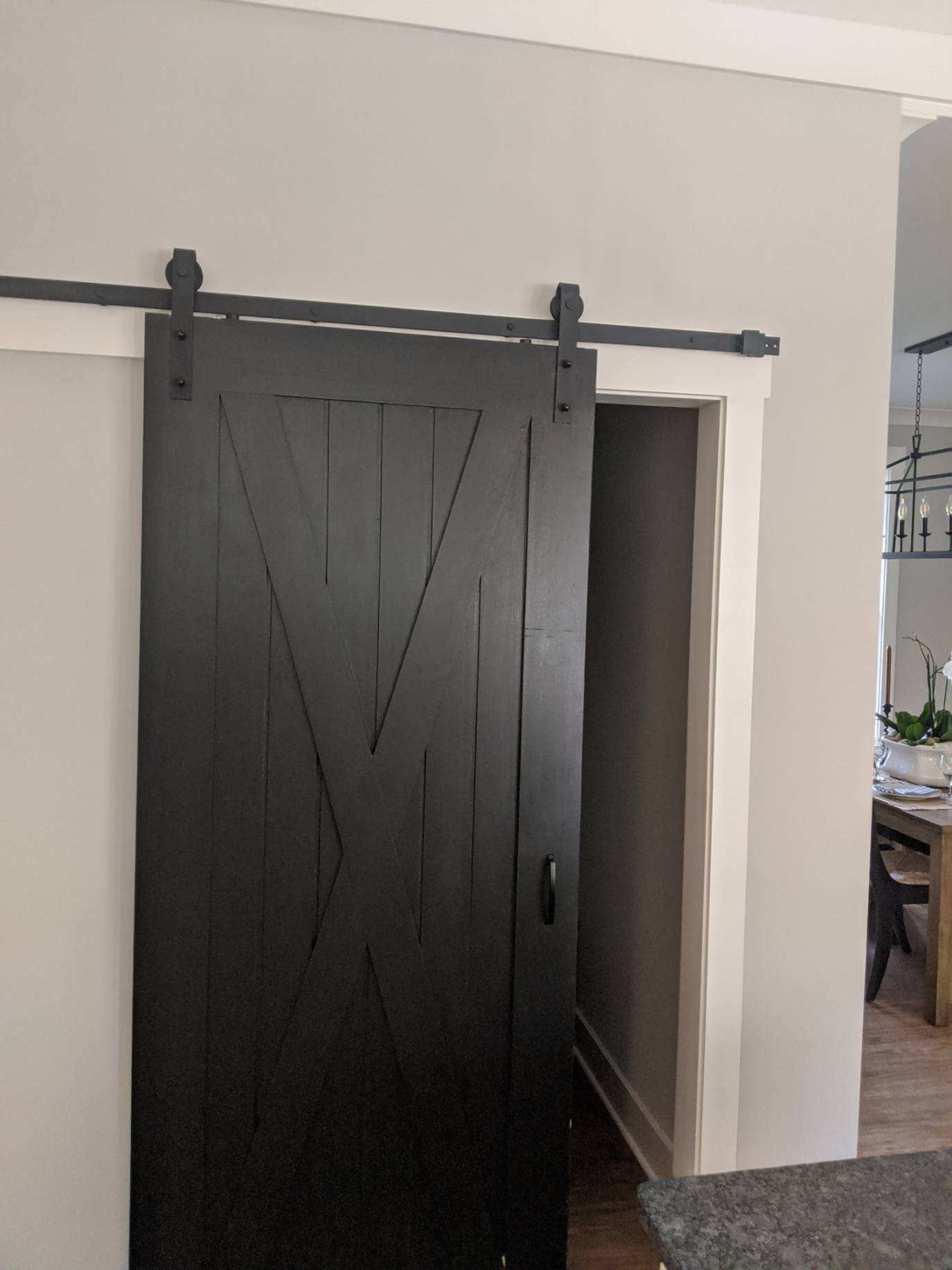 Black barn door partially open, mounted on a black track on a gray wall, doorway to a dining room.