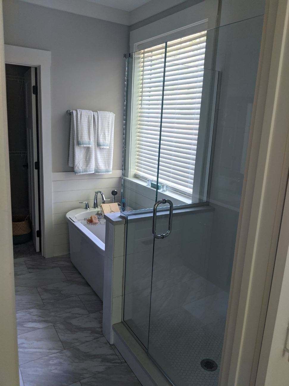 Bathroom with a glass shower, tub, window with blinds, white towels, and gray tile.