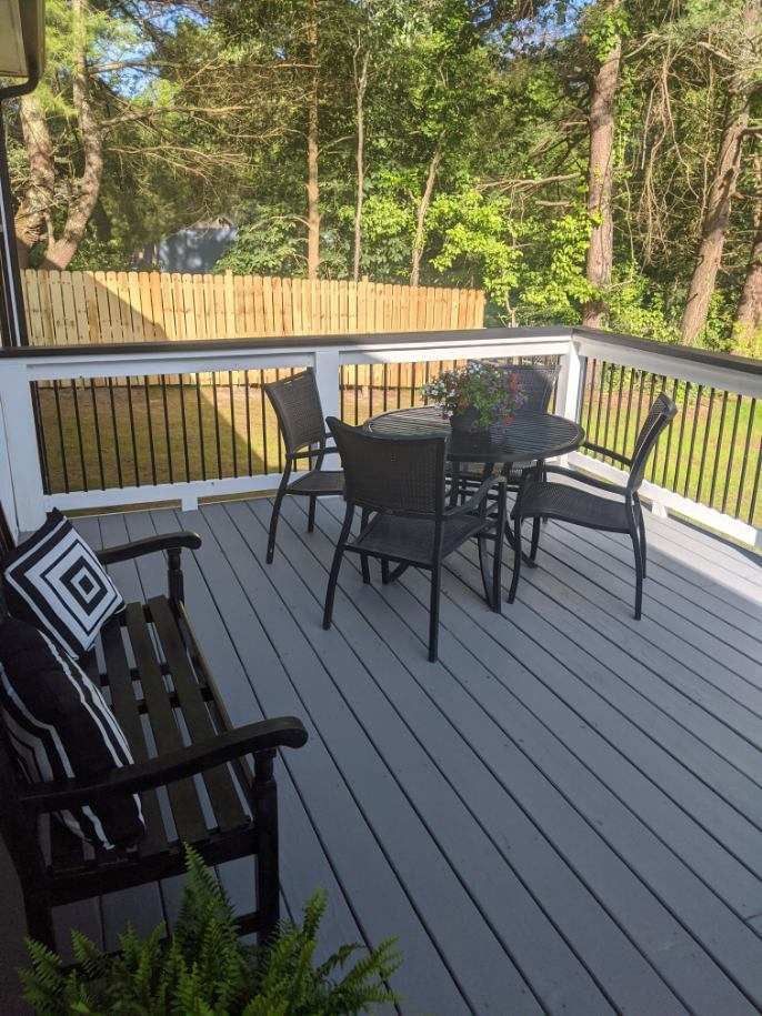 Deck with gray flooring, black furniture, and a wooden fence in the background, surrounded by trees.