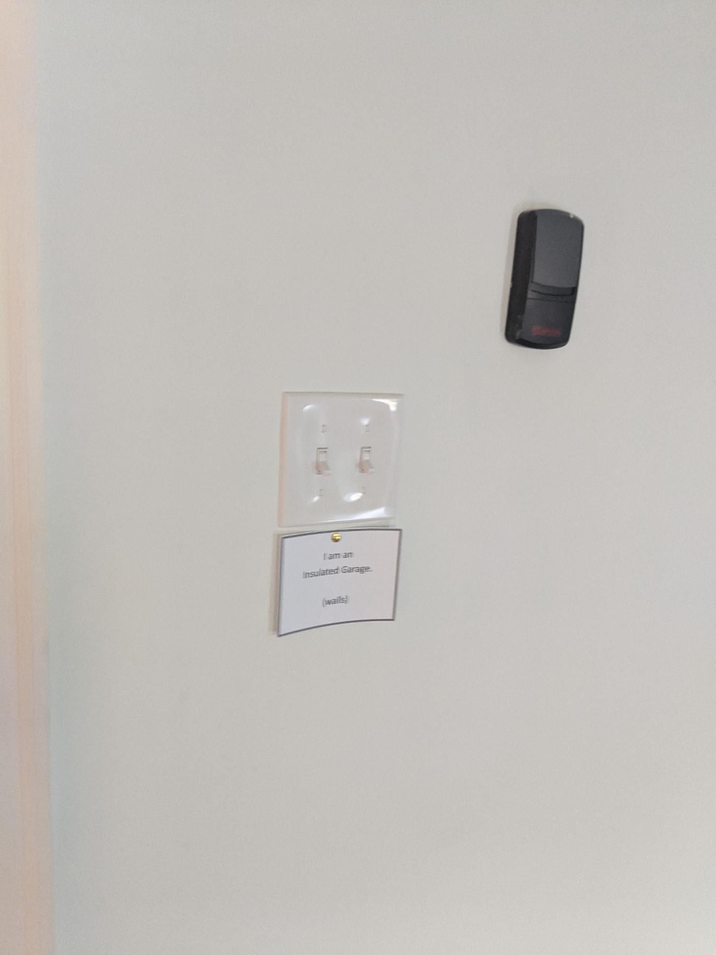 White wall with a light switch plate holding a note, and a black box mounted above.