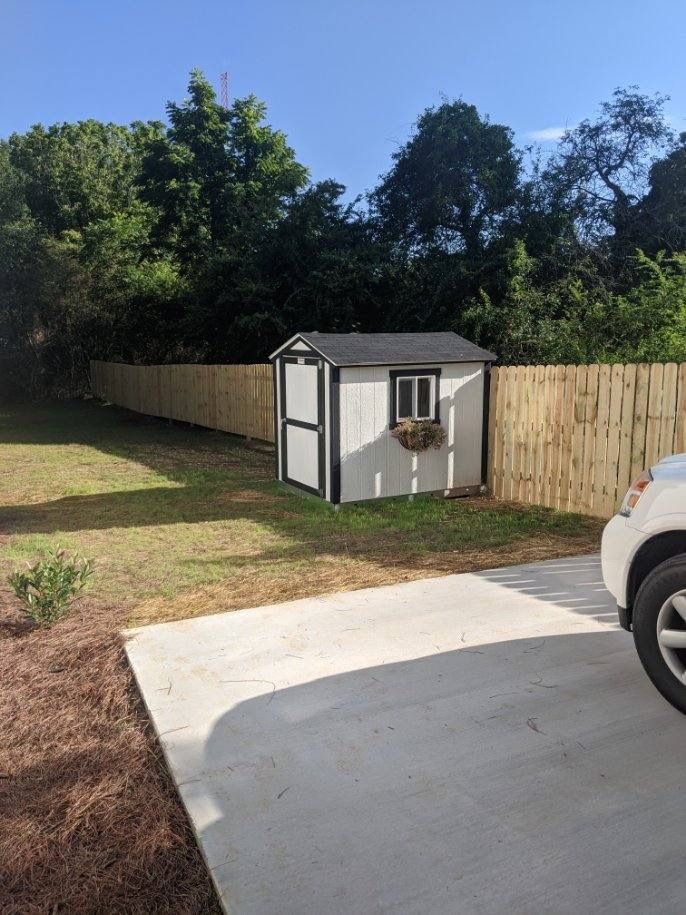 White shed with black trim and window box, set in a fenced backyard next to a concrete pad and a car.