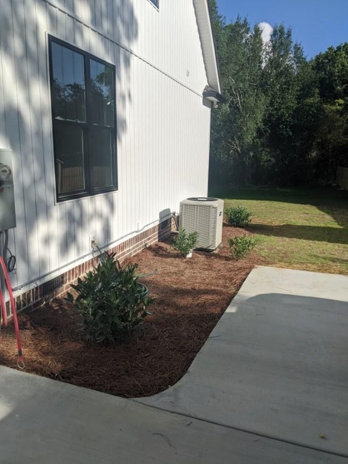White house exterior with black-framed window, an AC unit, and mulch-covered landscaping beside a concrete patio.