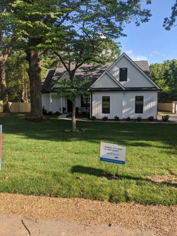 White house with black trim, set in front of a large tree. Green grass and blue sky.