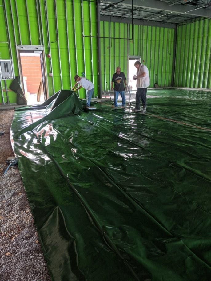 Three people installing a large green tarp on a wet concrete floor in a green-walled building.