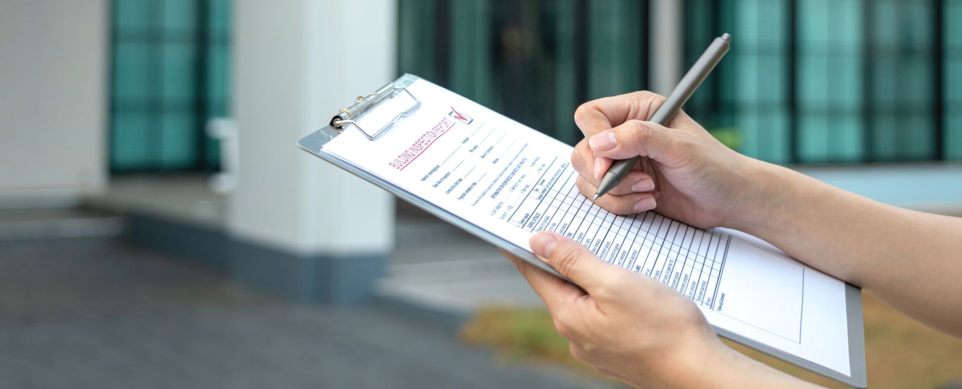 A person's hands hold a clipboard with a form and write with a pen in front of a house.