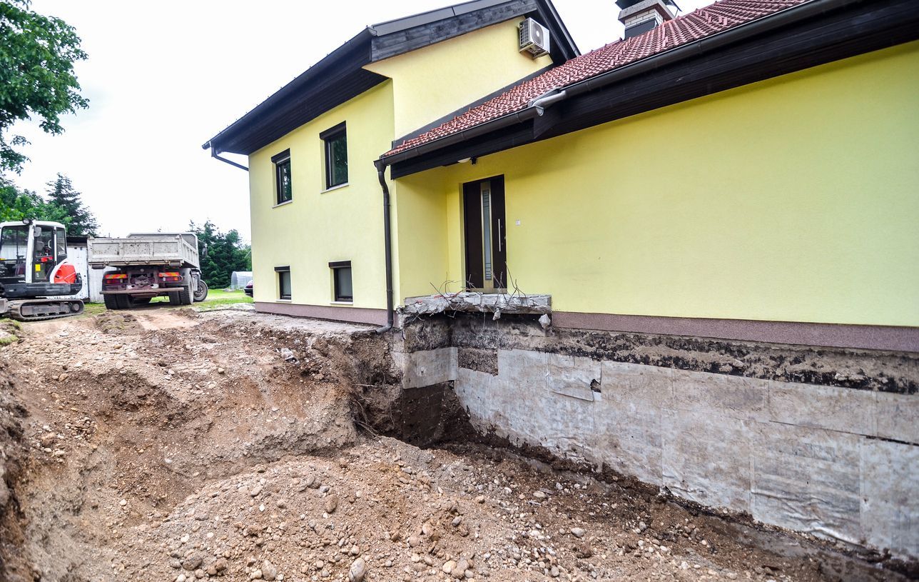 House with exposed foundation undergoing excavation. Yellow exterior, brown roof, with construction vehicles.