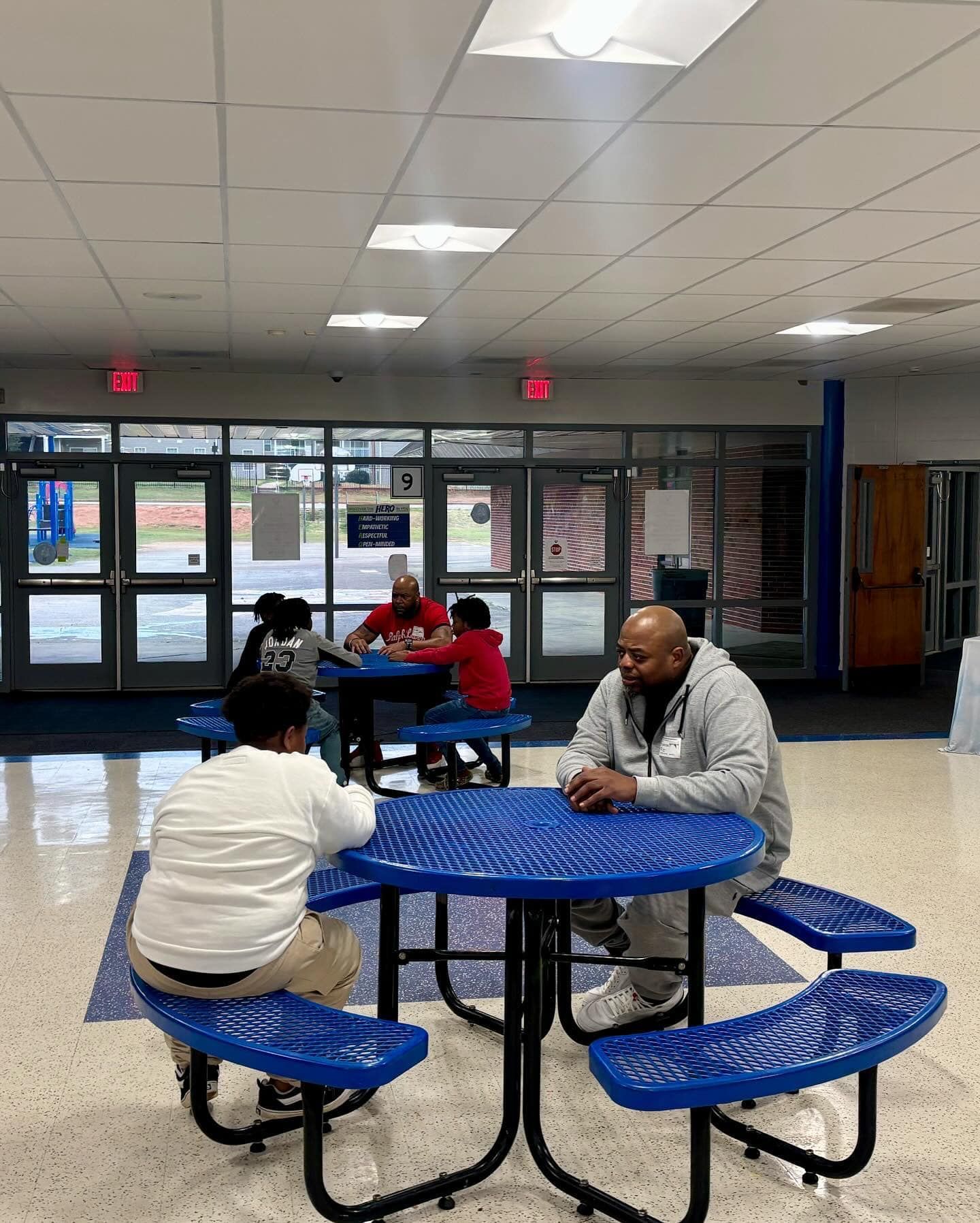 People sitting at blue picnic tables in a school hallway. Two adults, three children, a large window visible.