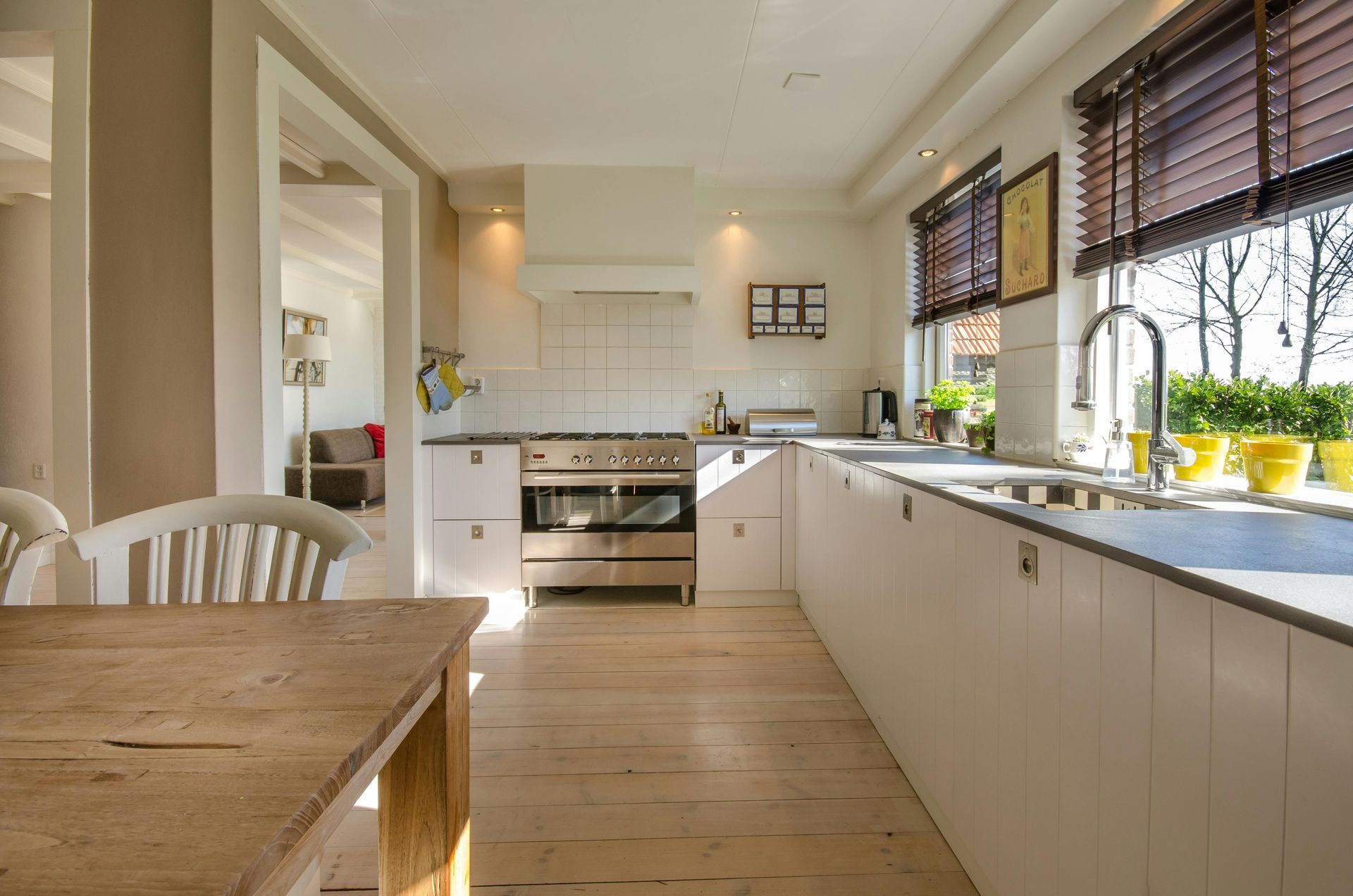 Bright white kitchen with wooden table, long countertop, stove, and sunlit windows with blinds.