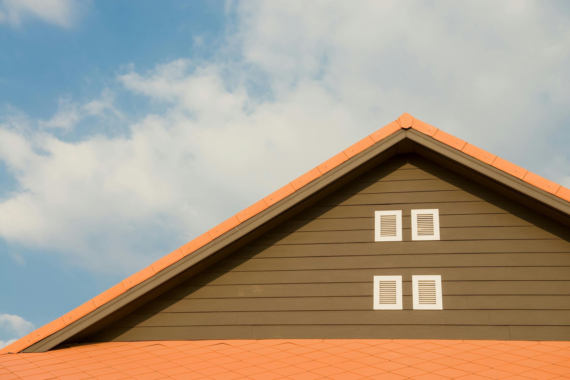 House gable with brown siding, white vent windows, and orange roof trim against a cloudy blue sky