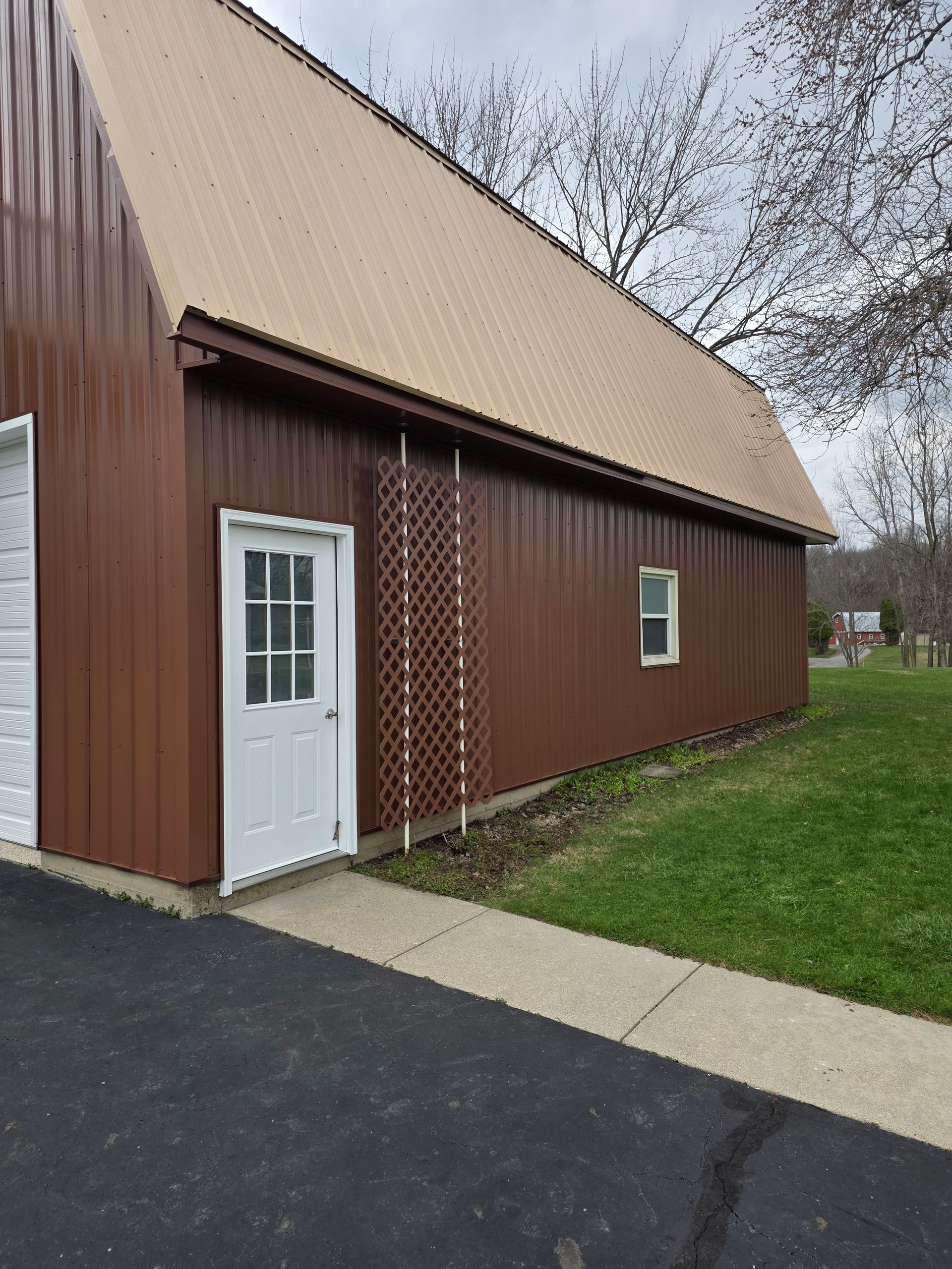 A side view of a brown metal-sided building with a white door, a small window, a wooden trellis, and a paved walkway.