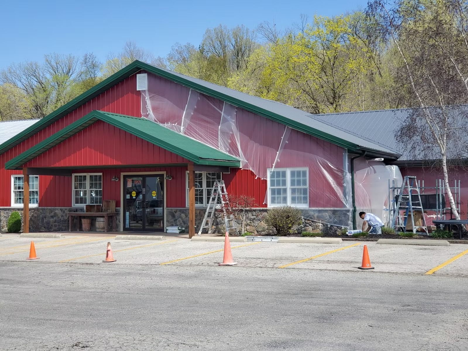 A red building with a green roof undergoing exterior renovations with plastic sheeting and ladders in a gravel lot.