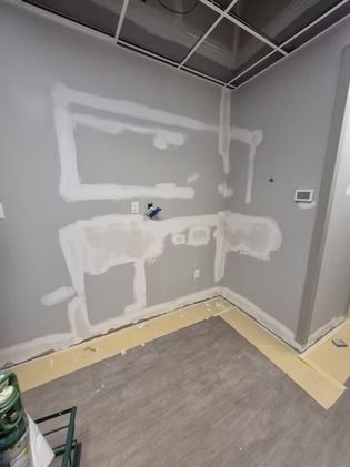 A room under renovation with gray walls, white drywall compound patches, exposed electrical wires, and flooring tape.