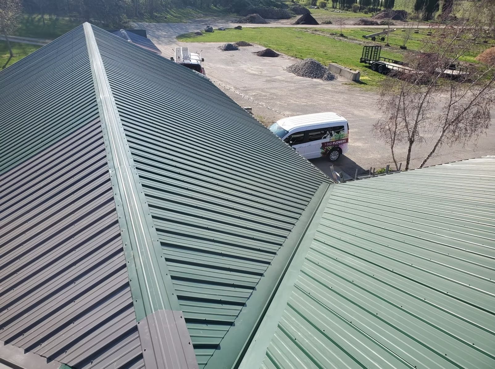 A high-angle view of a green metal roof with a central valley meeting two roof sections, with a van parked below.