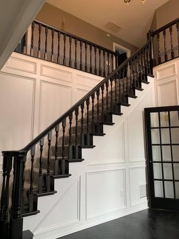 A grand foyer features a black wooden staircase, white wainscoting, and a matching black French door on dark wood floors.