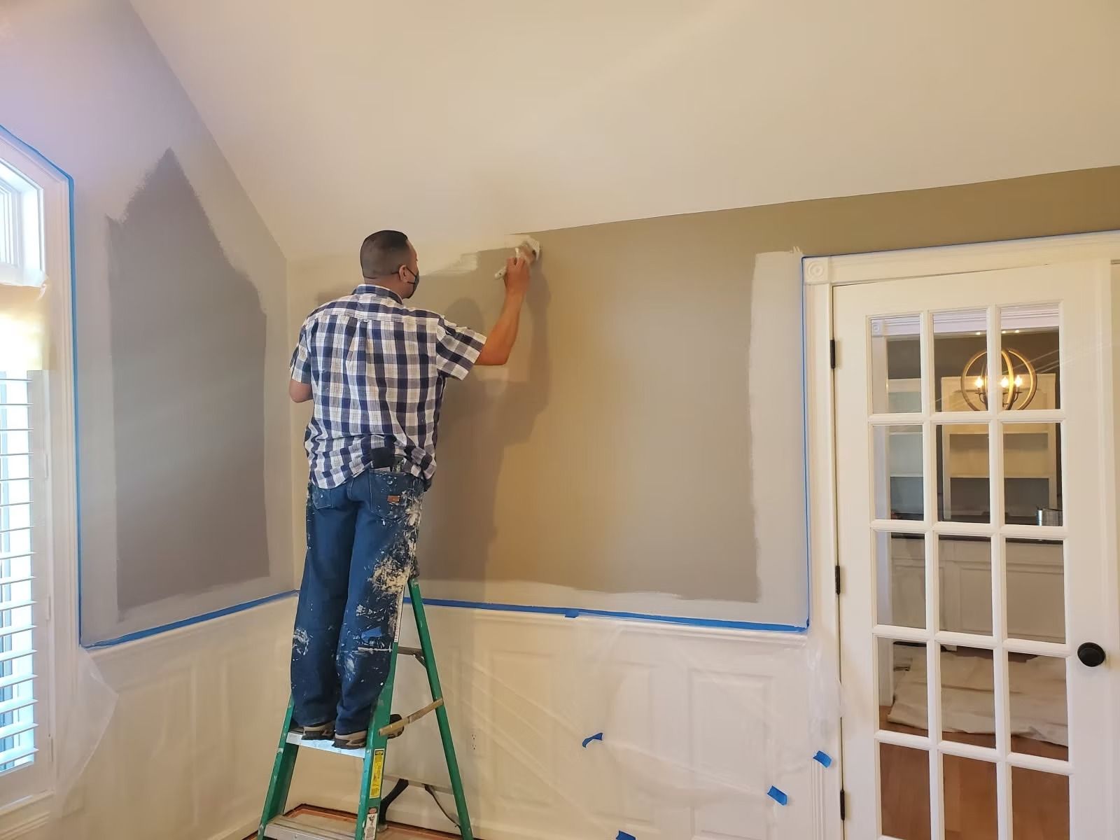 A person stands on a ladder, painting a section of a beige wall above white wainscoting in a room with a glass-paneled door.