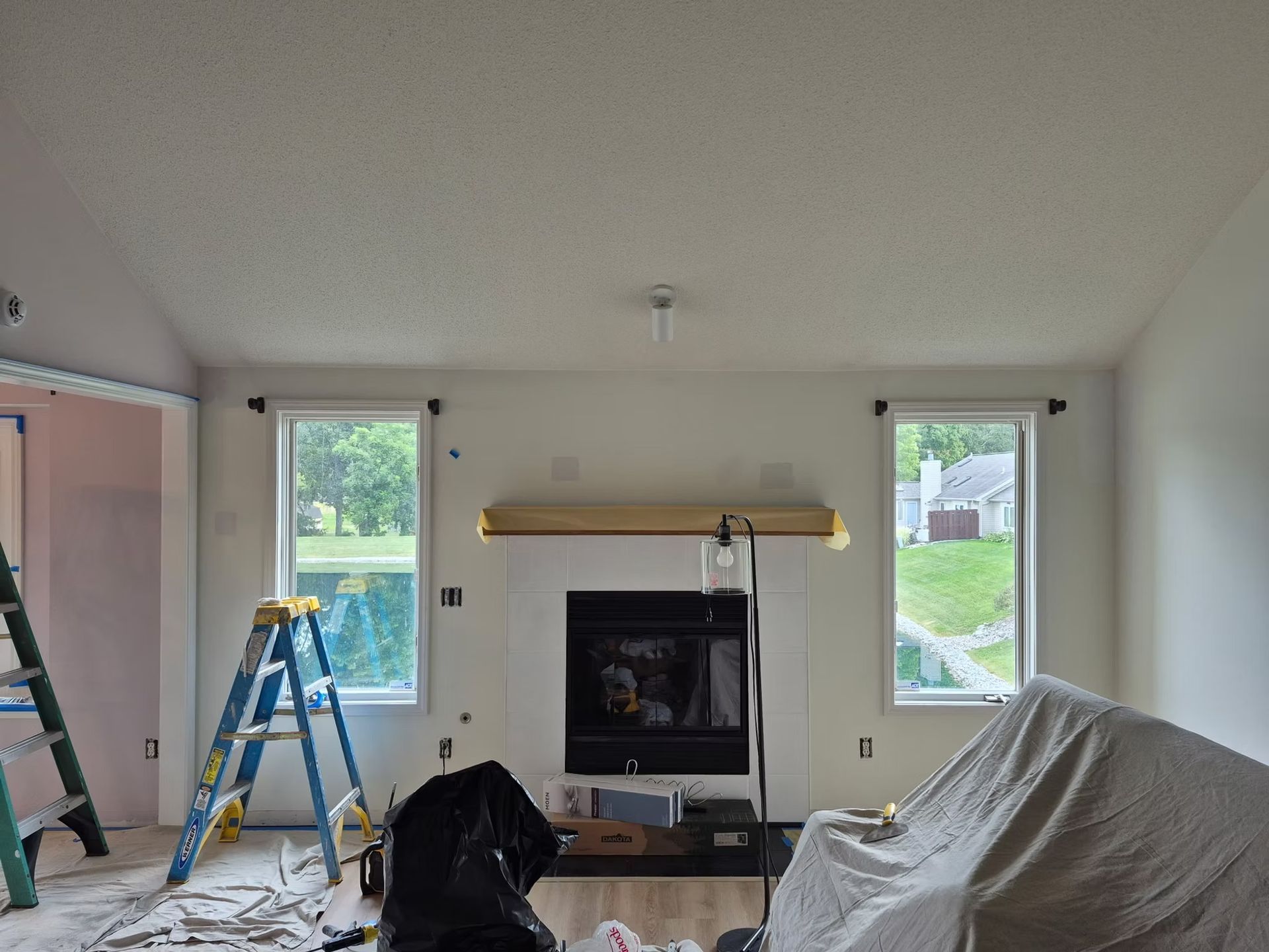 A room under renovation featuring a fireplace, two windows, a ladder, and furniture covered with a drop cloth.