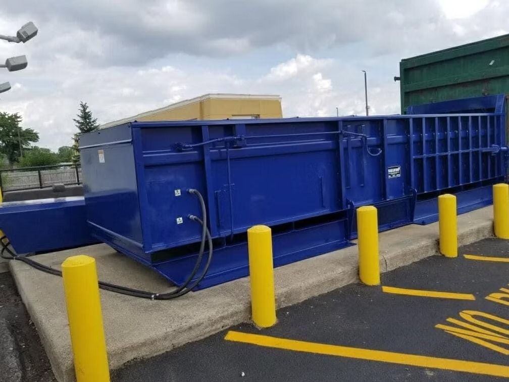 A large, rectangular blue industrial trash compactor sits on a concrete pad with yellow safety bollards in front.