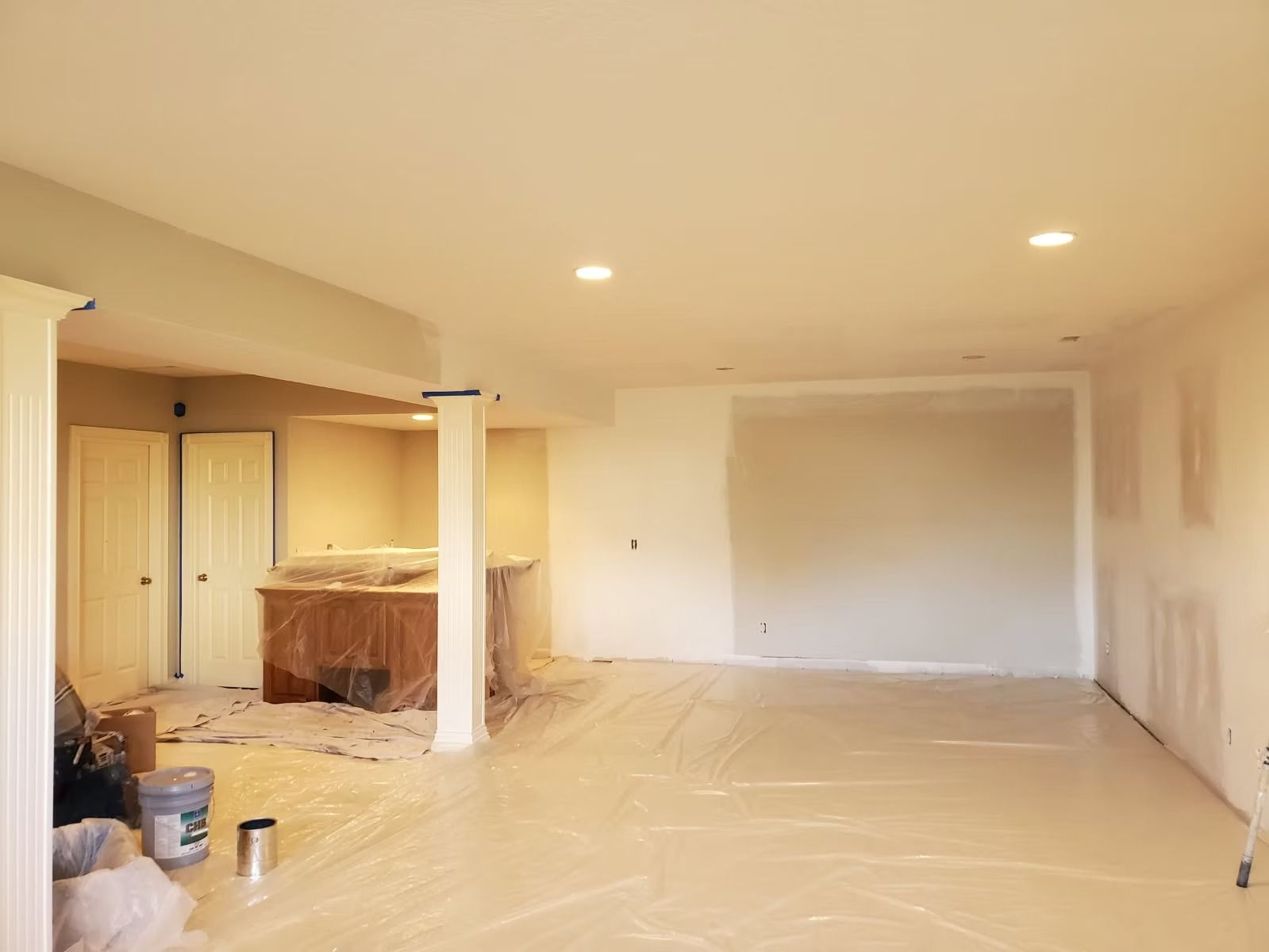 An unfinished interior room with neutral walls, white pillars, recessed lighting, and a plastic-covered floor.