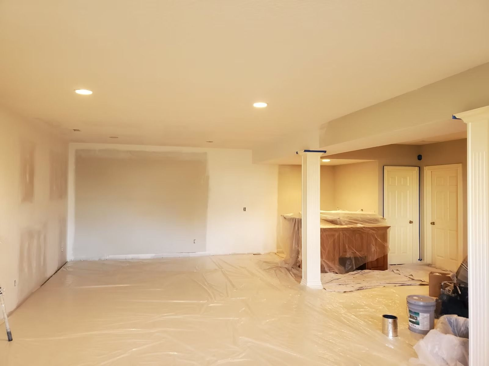 A room under renovation with taped drywall, plastic floor protection, white columns, and recessed lighting.