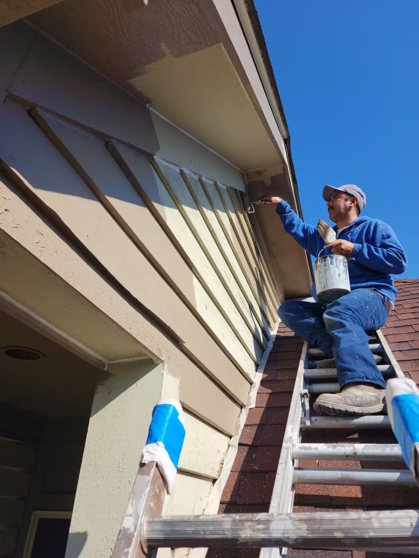 A person in a blue hoodie and jeans on a ladder, painting the exterior wall of a house under a clear blue sky.