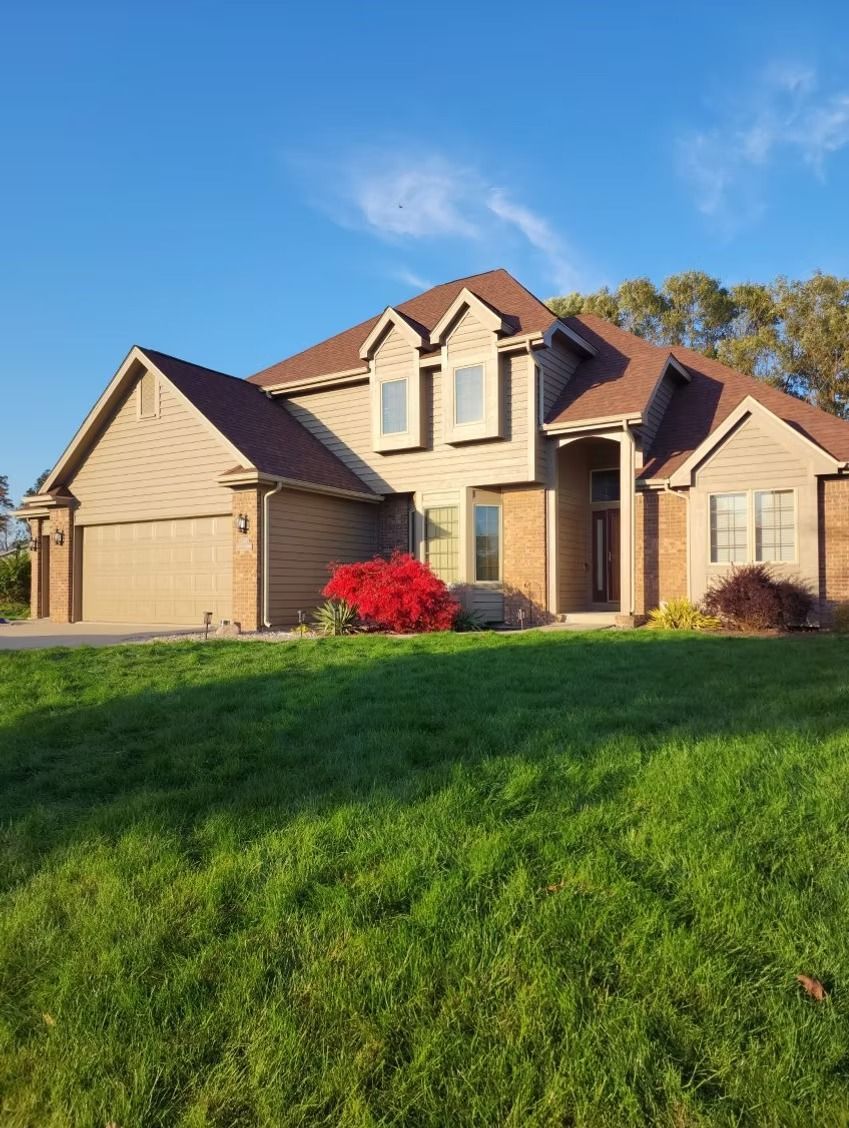 A two-story suburban house with beige siding, a brown roof, and a large front lawn under a clear blue sky.