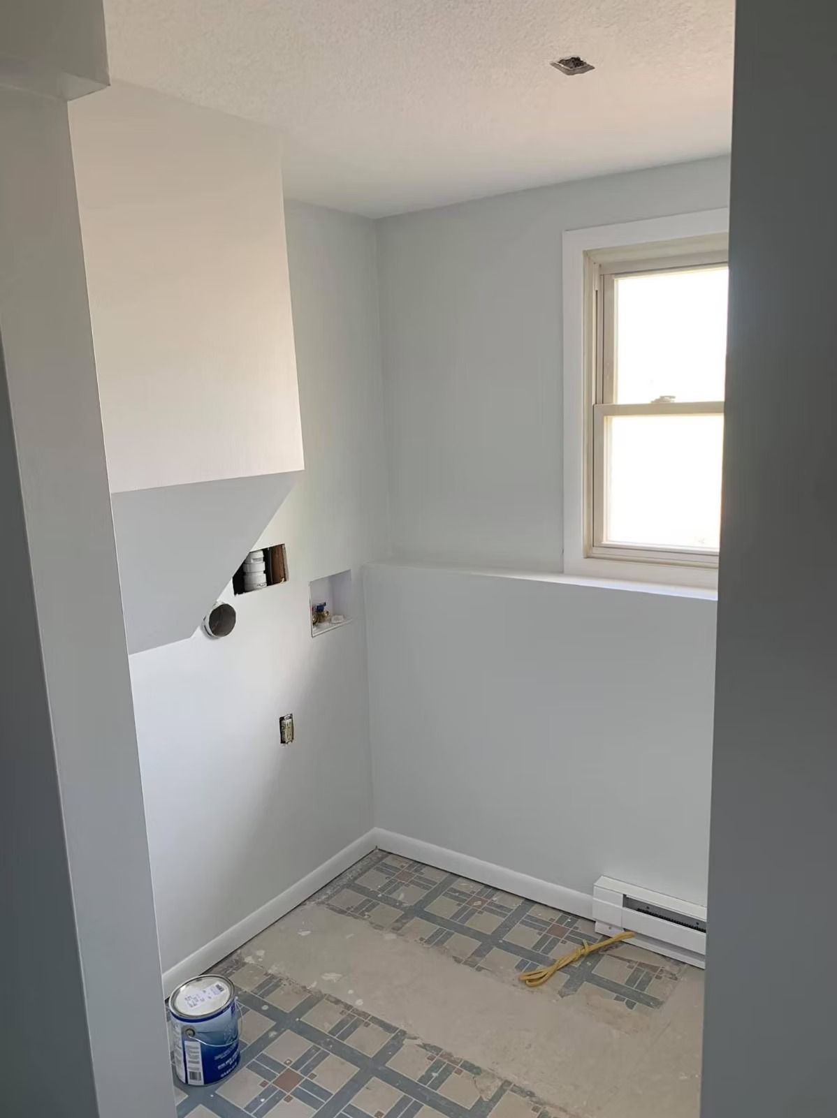 A small, empty laundry room with light gray walls, a window, unfinished plumbing hookups, and patterned tile flooring.