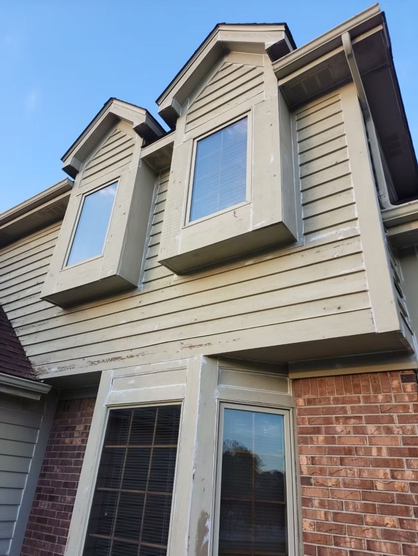 Exterior view of a two-story house with tan siding, brick accents, and two prominent upper-level bay windows.
