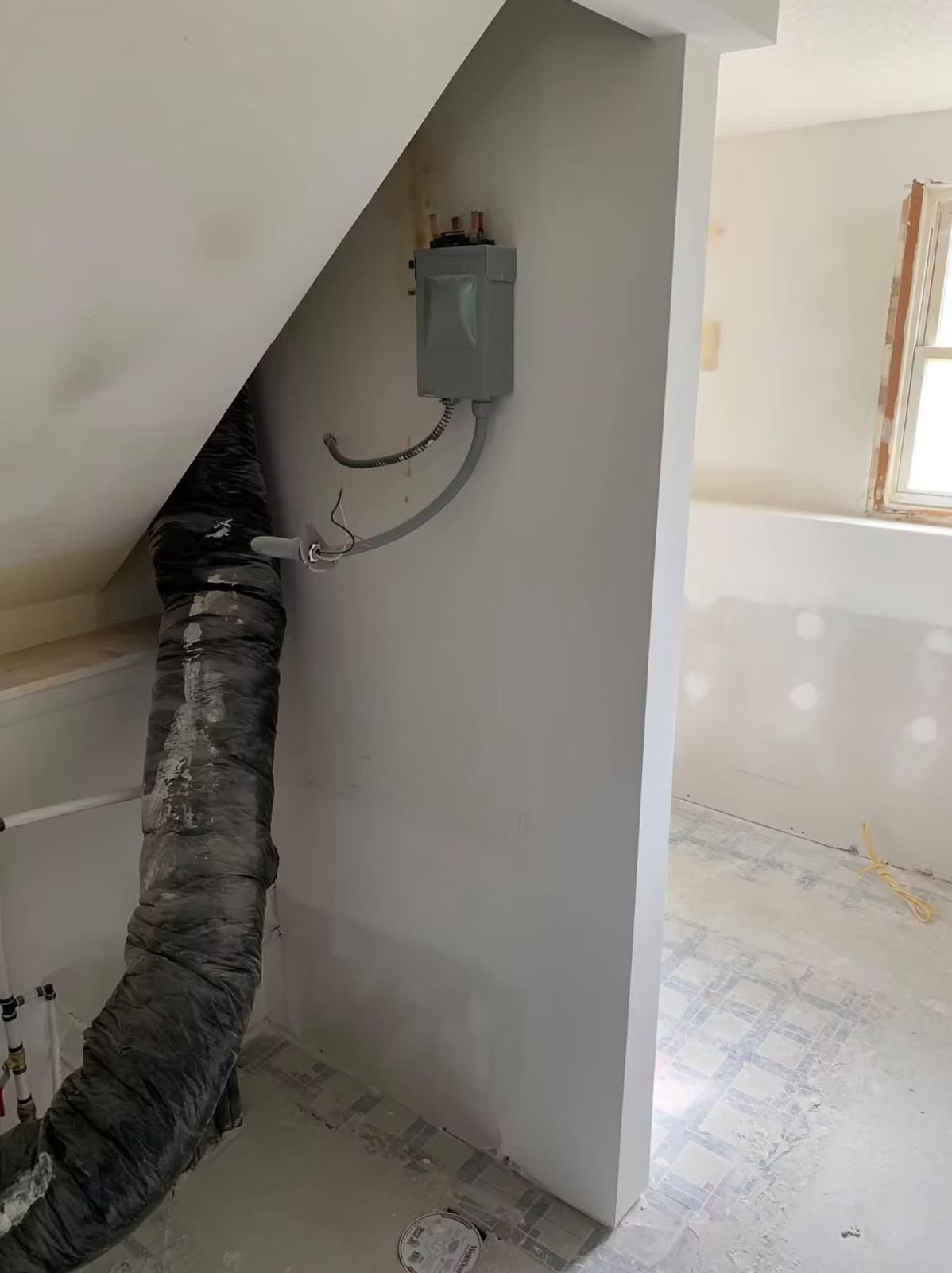 An electrical disconnect box mounted on a white wall under a slanted ceiling, next to a vertical flexible duct pipe.