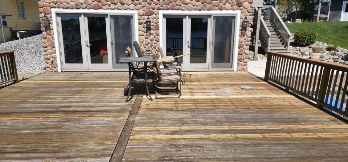 A wood deck with uneven coloring and wear, featuring a small bistro set in front of a stone house with sliding glass doors.