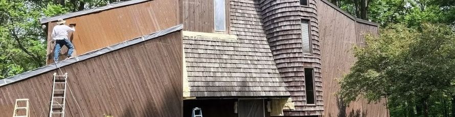 A person stands on the sloped roof of a brick house undergoing exterior construction and repair.