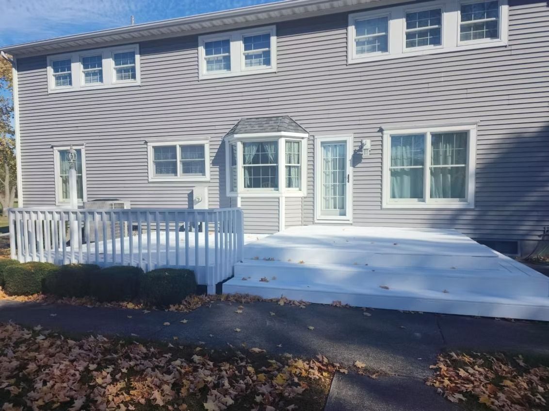 A two-story house with gray siding features a large, freshly painted white deck and railings in a yard with fallen leaves.