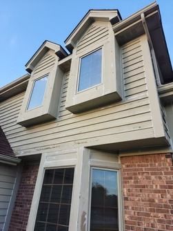 A low-angle view of a beige, two-story residential house exterior featuring a brick base and two dormer windows above.