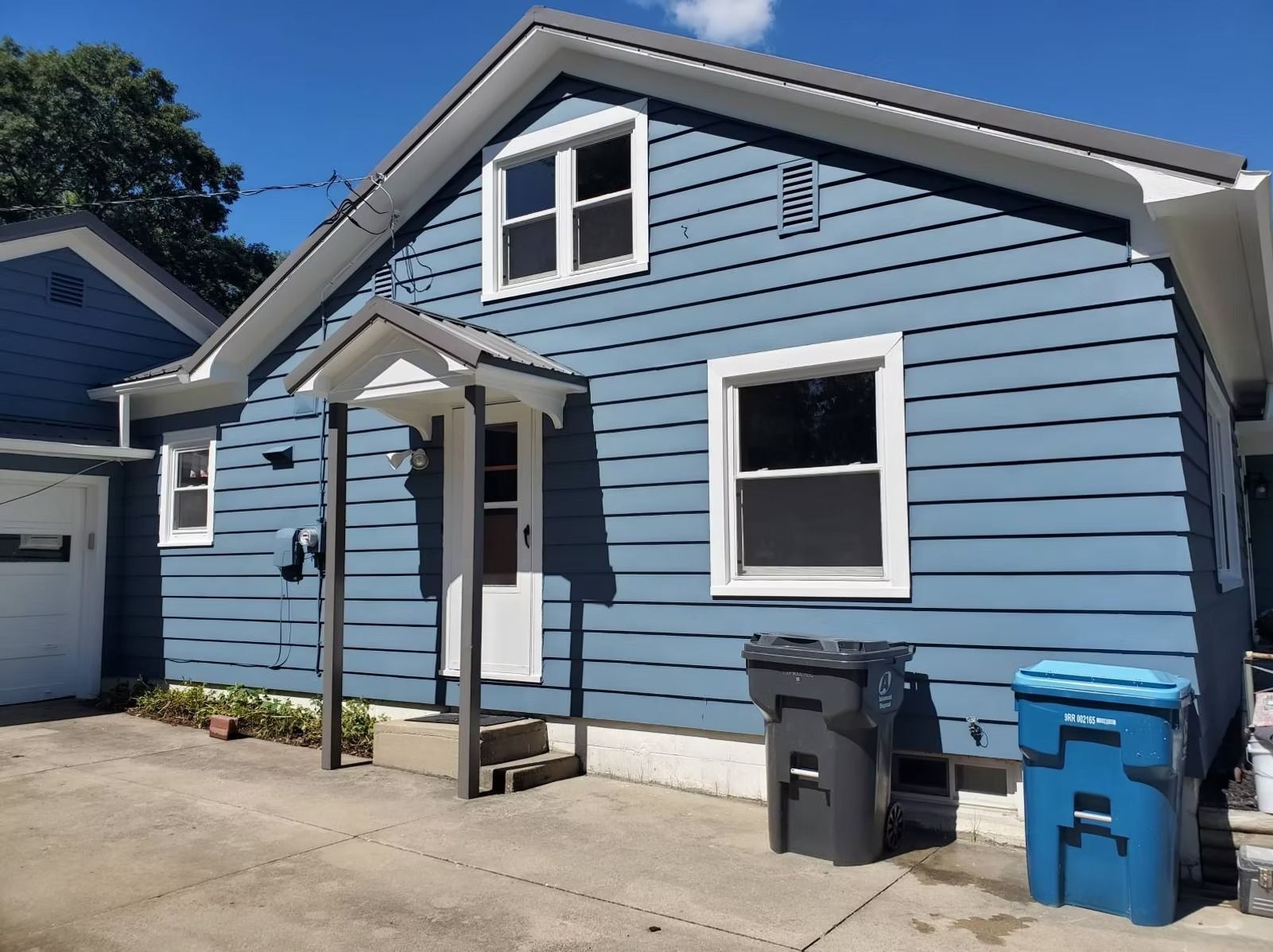 Blue house with a small front porch, a gabled roof, and two trash cans on a concrete driveway in daylight.
