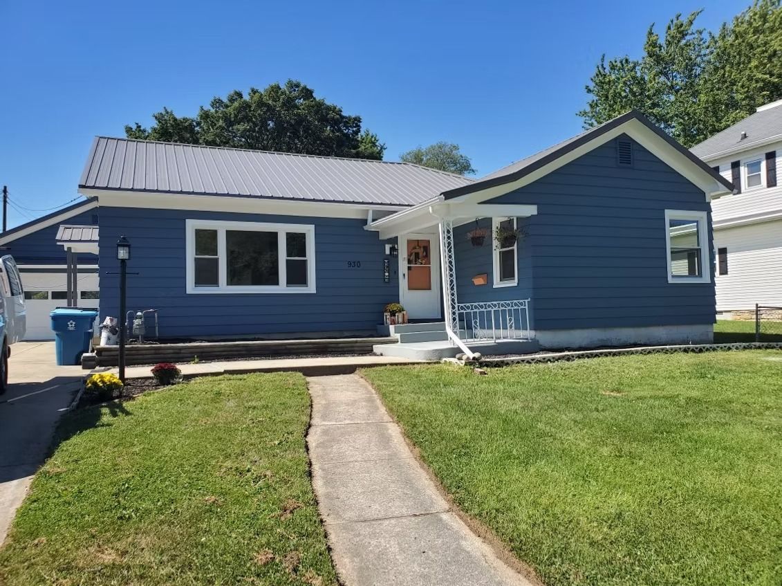 A blue, single-story house with a white trim, metal roof, and a paved walkway leading to the front entrance.