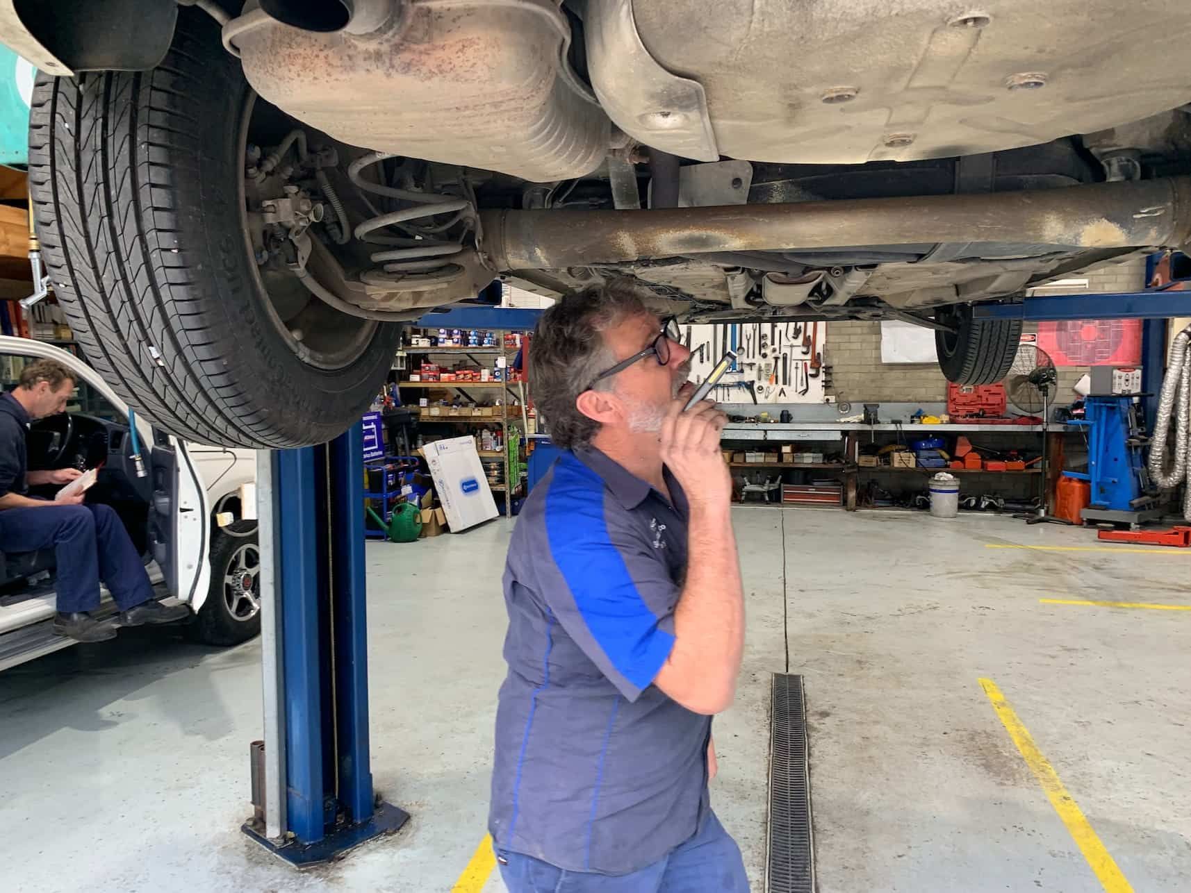 A Man is Standing Under a Car on a Lift in a Garage — Town & Country Removals In Taree, NSW