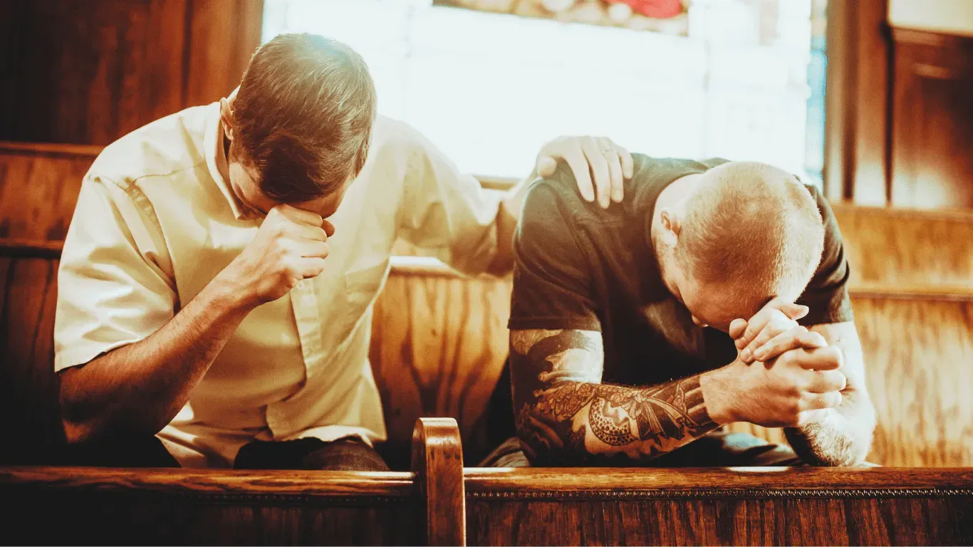 Two people kneel in a church pew, heads bowed in prayer with hands clasped.