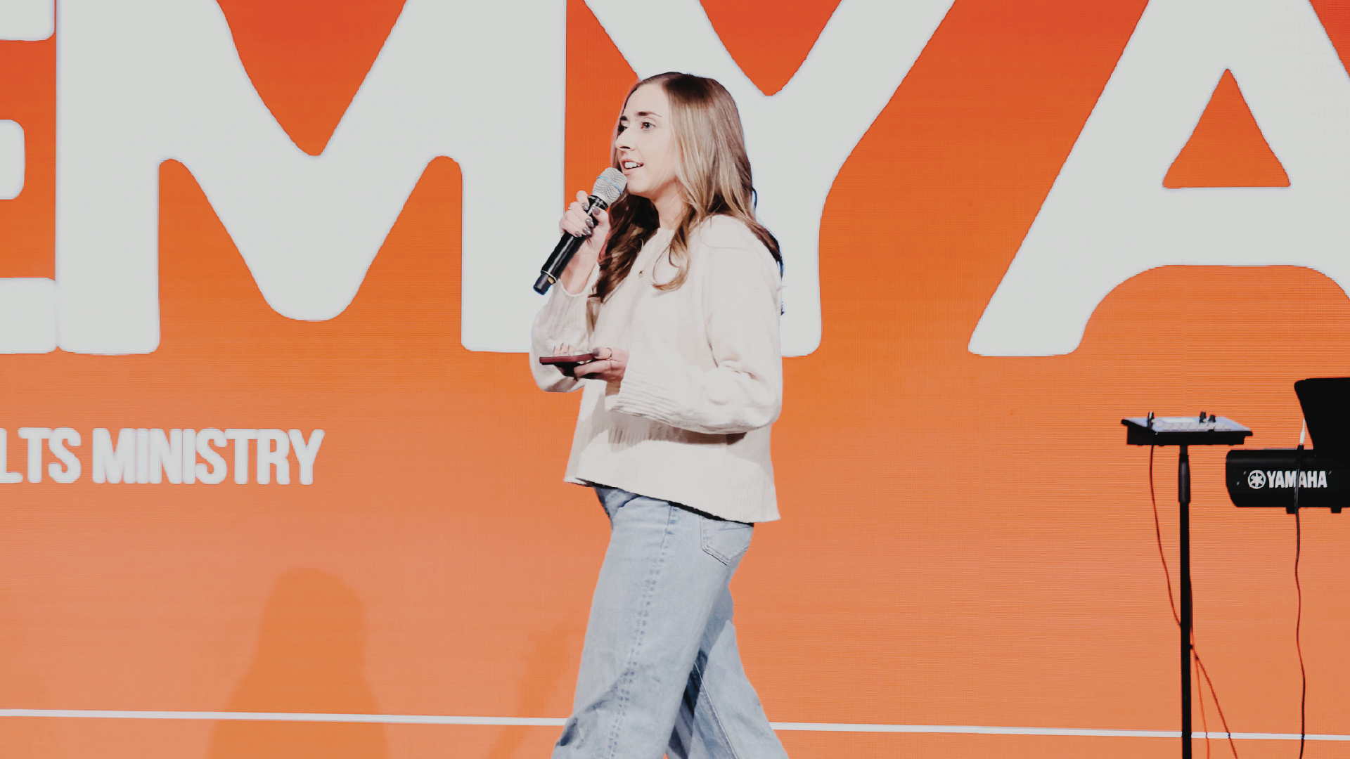Woman speaking into a microphone on an orange stage backdrop at a ministry event